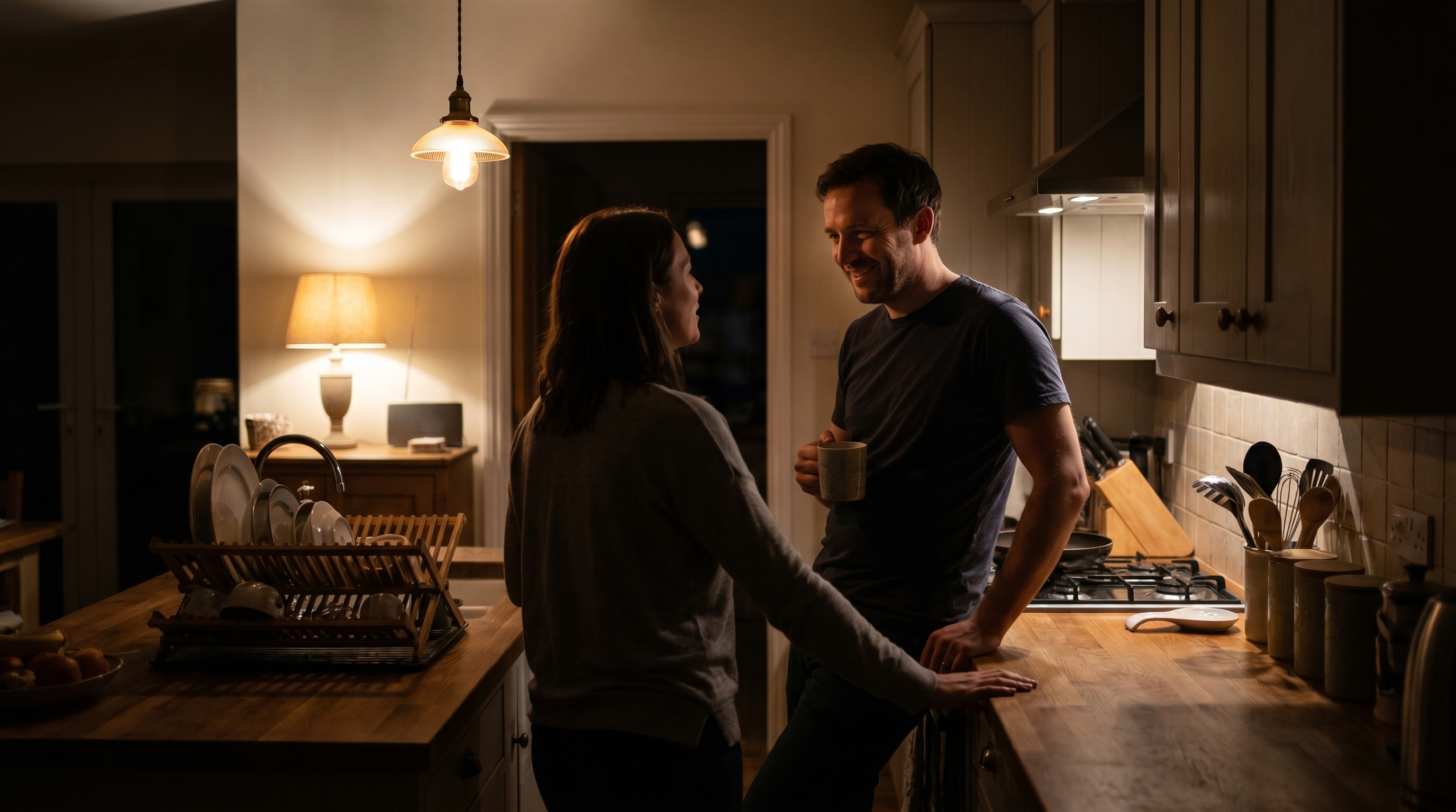 A couple in a low-lit kitchen, close together, mid-conversation over a quiet evening
