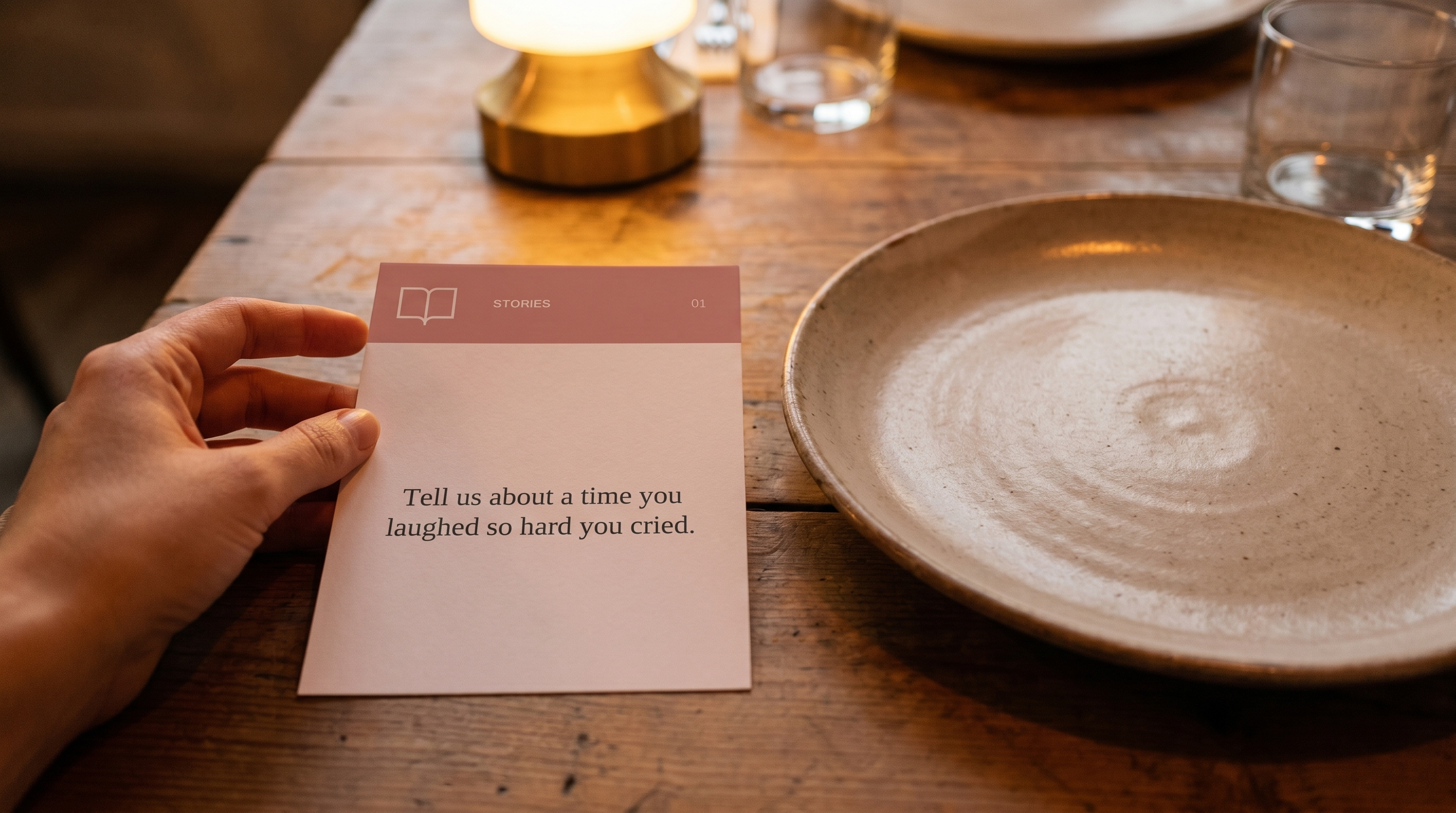 Hands placing a printed conversation card next to a plate on a dinner table, warm evening light